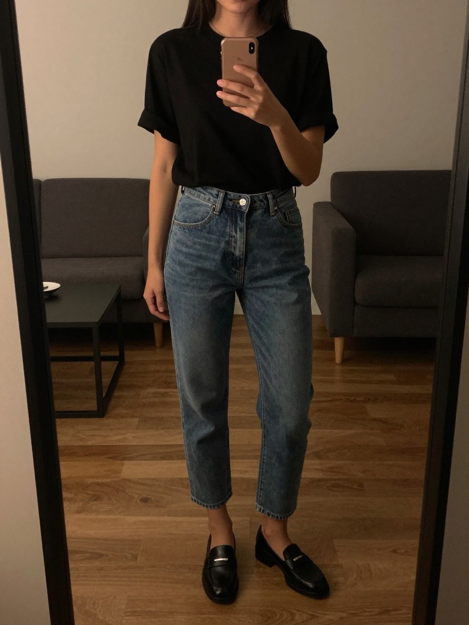 iPhone mirror selfie of a woman in cropped jeans and loafers, holding her phone at chest level with one hand behind her back, cropped at NECK so face doesn't show, in a city apartment, warm moody lighting, iPhone photo quality.