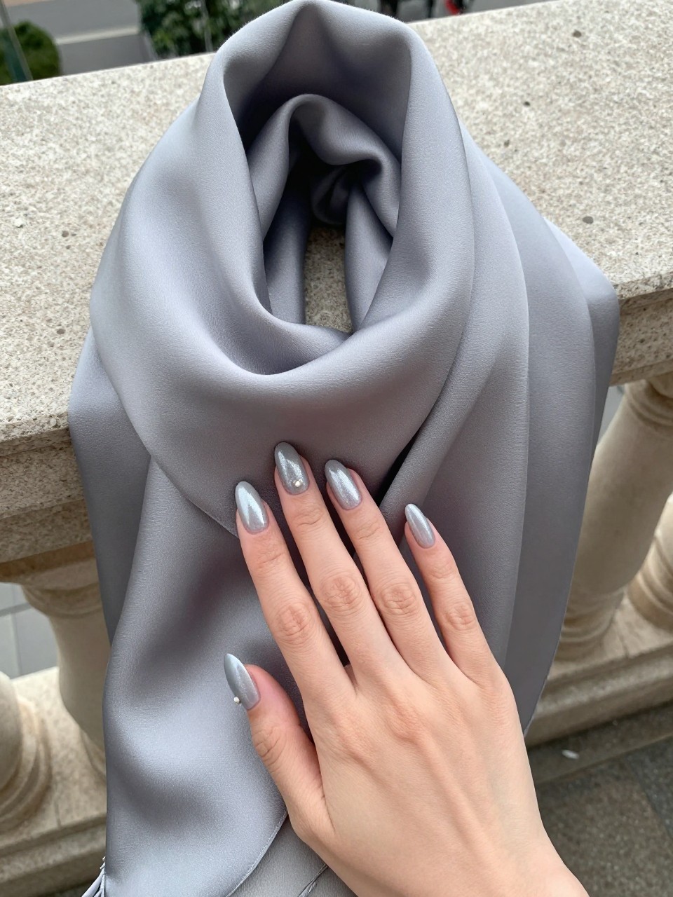 Top-down view of hands on a sleek, grey silk scarf on a stone balcony. The almond nails are a soft grey with a delicate pearl shimmer. The diffuse, overcast daylight makes the subtle sheen glow.