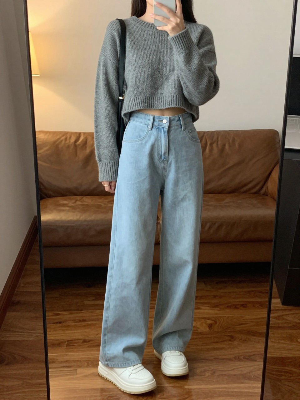 iPhone mirror selfie of a woman in a grey cropped sweater and light blue wide leg pants with chunky white sneakers, relaxed stance holding phone, cropped at NECK so face doesn't show, in a cozy city apartment, warm moody lighting, iPhone photo quality.