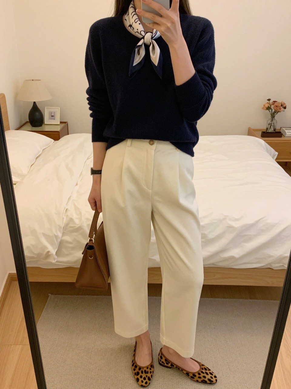 iPhone mirror selfie of a woman in a navy jumper with cream trousers and leopard flats, adjusting a silk scarf tied at her neck, cropped at NECK so face doesn't show, in a feminine-themed bedroom, warm moody lighting, iPhone photo quality.
