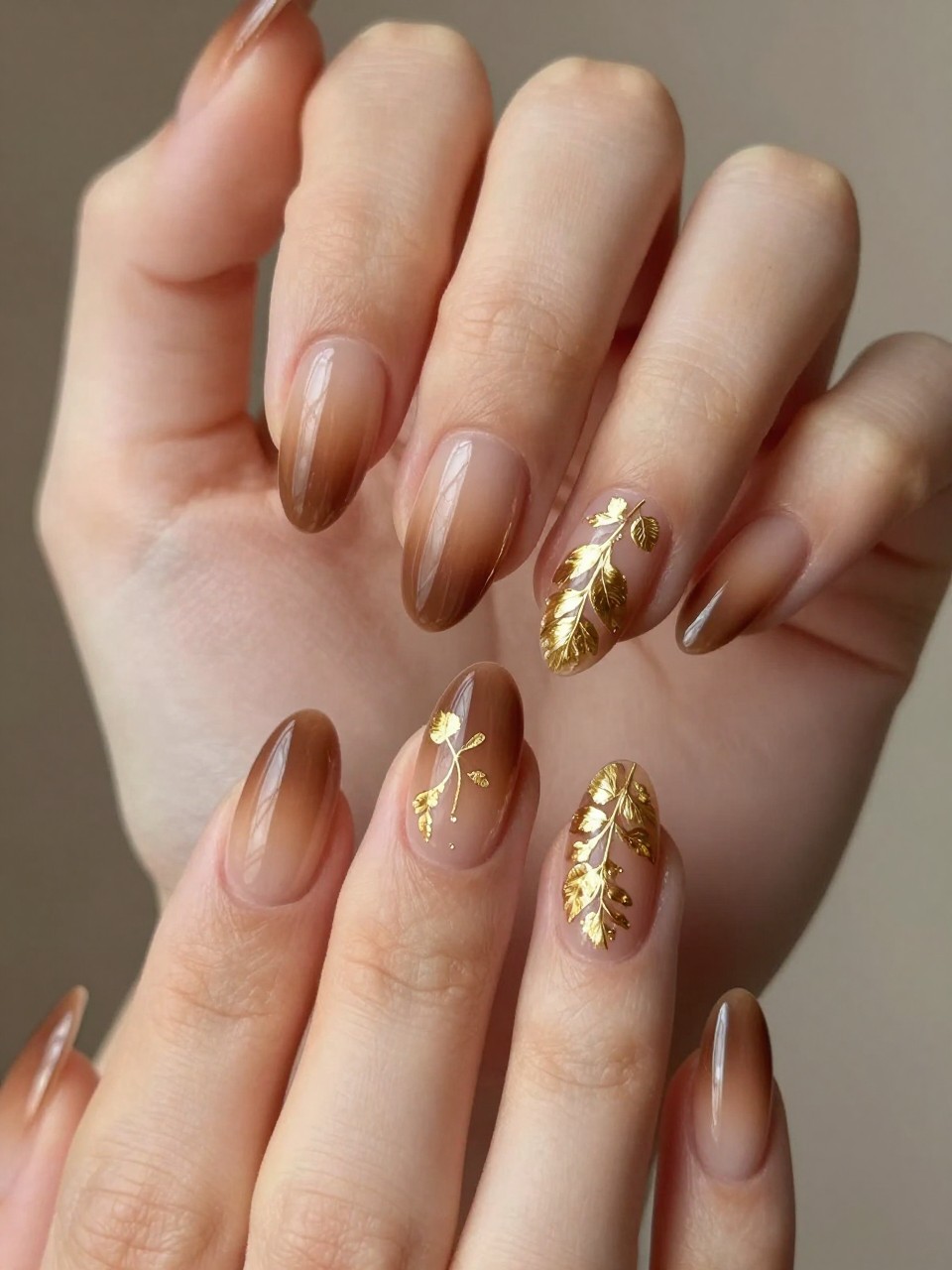 Close-up of two hands with light catching the gold details. The almond nails feature a smooth brown-to-tan ombré. Delicate gold leaf vines with small leaves trail across the gradient. The effect is warm and luxurious.