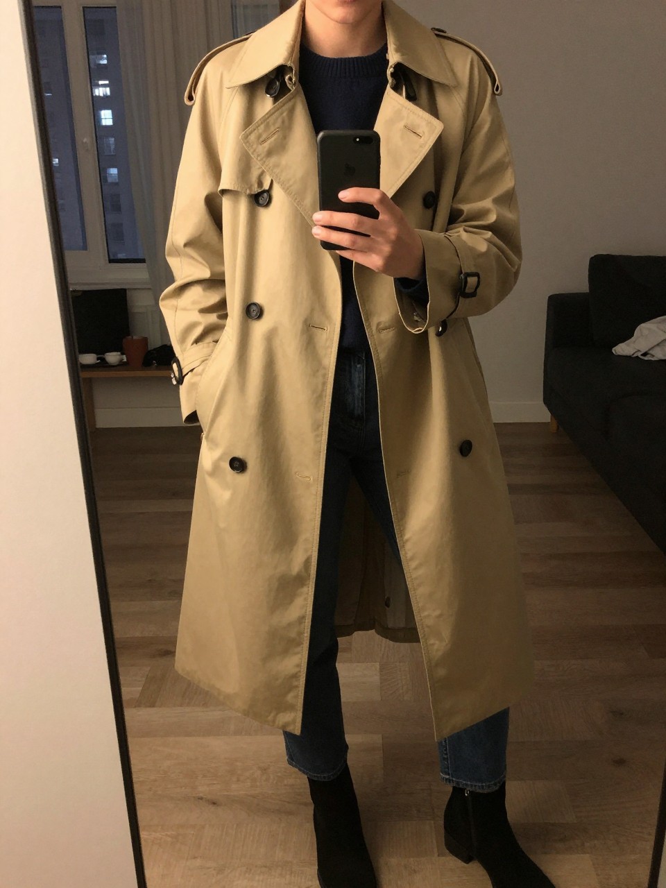 iPhone mirror selfie of a woman in a beige trench coat over a navy crewneck sweater and dark jeans with ankle boots, standing with phone at waist level, cropped at NECK so face doesn't show, in a city apartment, warm moody lighting, iPhone photo quality.