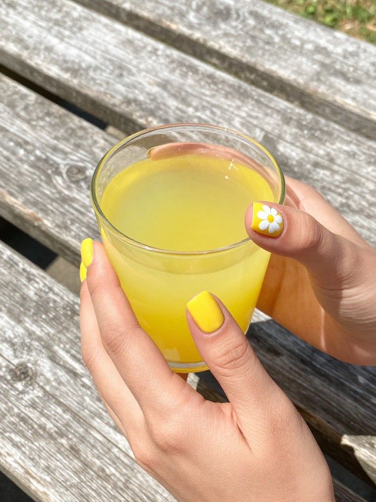 Overhead shot of hands holding a bright yellow lemonade. The nails are a short square shape, painted a glossy, cheerful yellow. Each thumb nail features a single, white five-petaled daisy with a tiny yellow dot center. The background is a rustic wooden picnic table in full sun.
