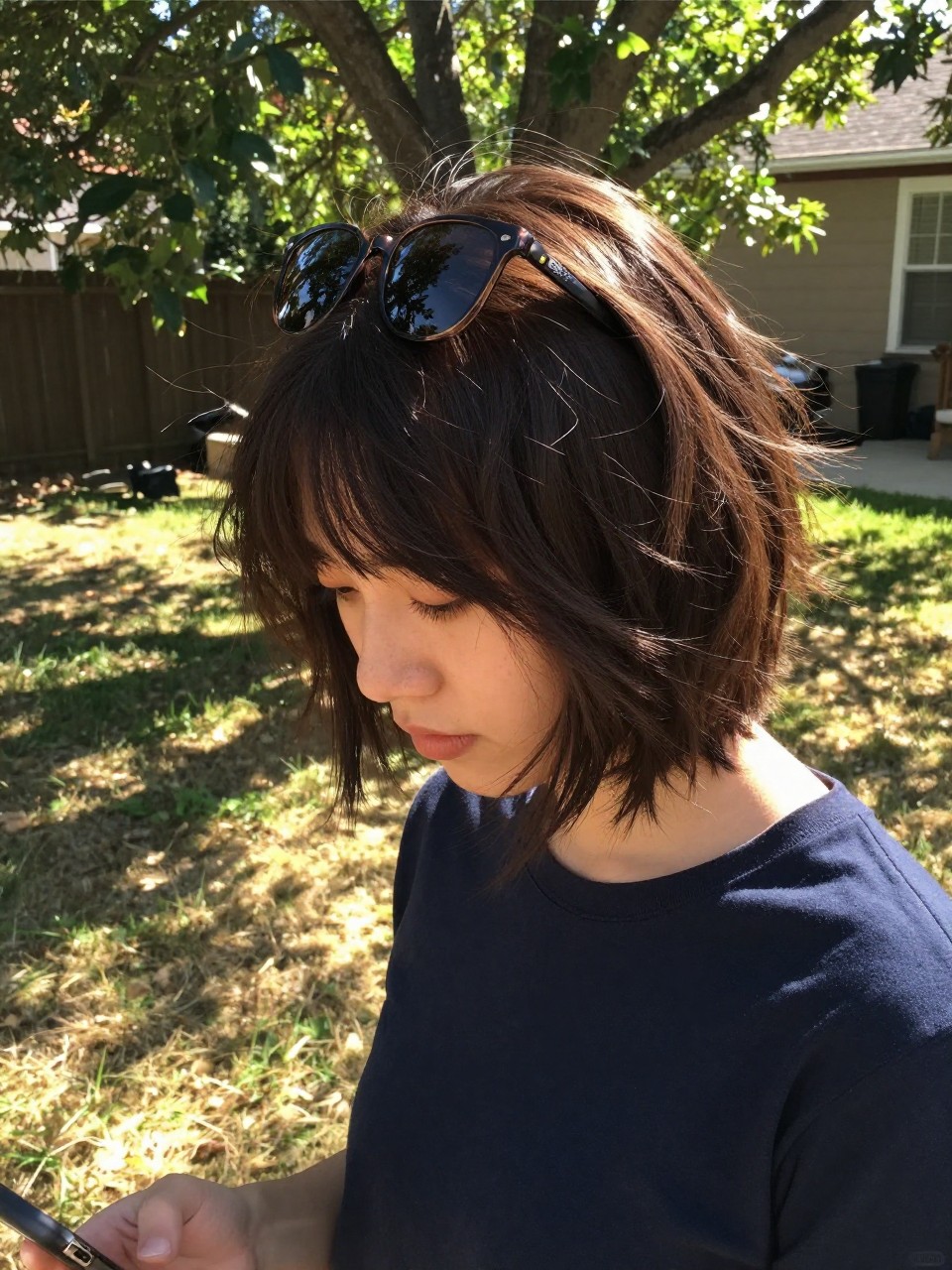 Photo of shaggy layered bob, looking down shot as if fixing hair, in a sunny backyard with patchy grass, dappled sunlight filtering through tree leaves, containing a pair of sunglasses perched on the head, iPhone photo quality.