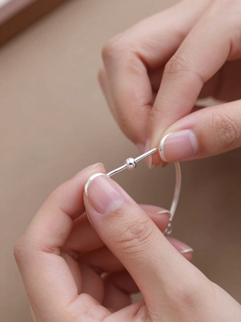 Close-up overhead view of hands fastening a delicate silver bracelet. The short, oval nails have a translucent pink base. A micro-thin line of metallic silver traces the very edge of each tip. The focus is sharp and detailed.