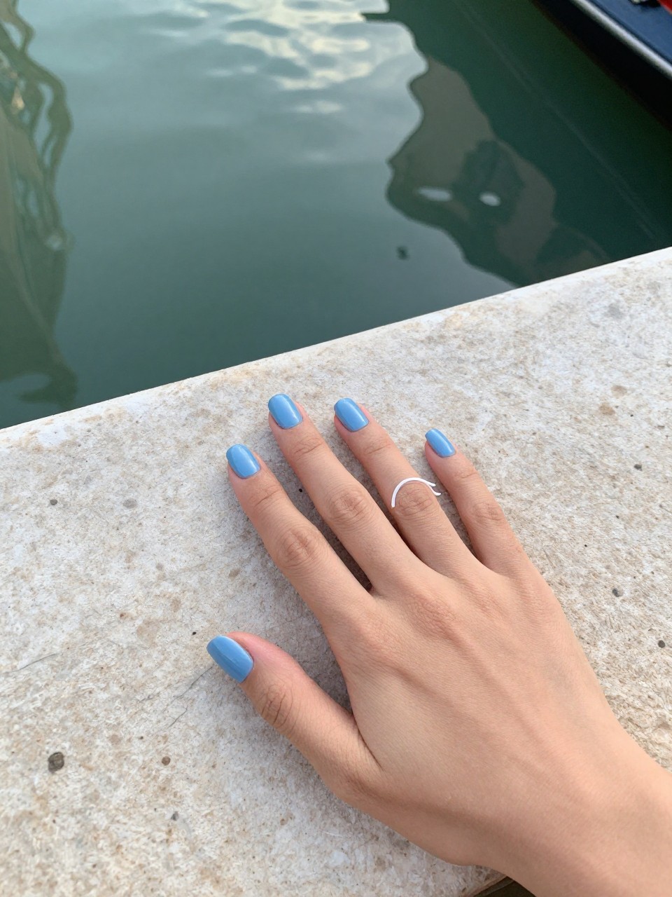Overhead view of hands resting on a stone windowsill overlooking a canal. The nails are a soft, glossy azure blue. A single, clean white gondola outline glides across each ring finger. The afternoon light is soft and reflective on the water.