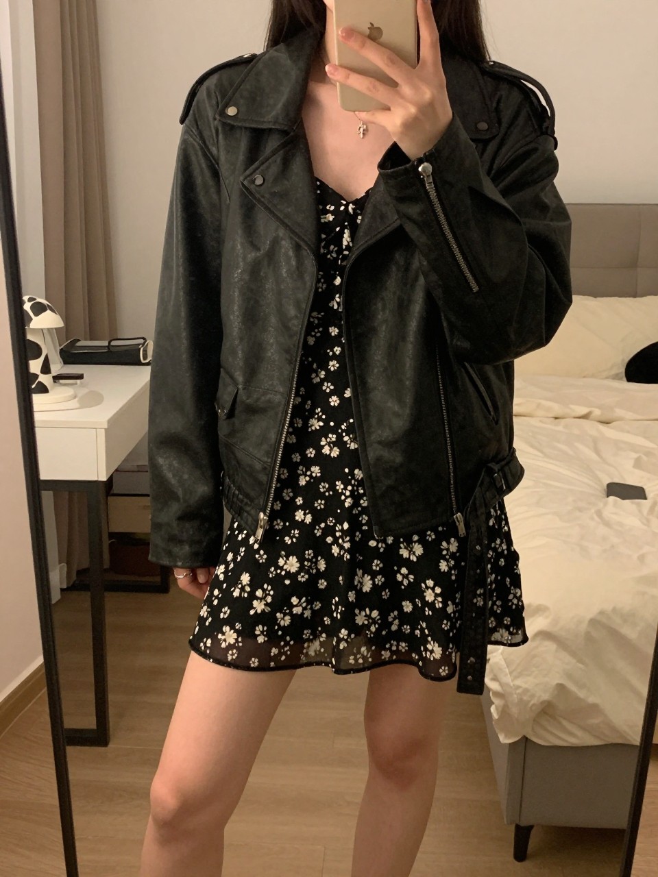 iPhone mirror selfie of a  woman wearing a black leather jacket over a floral dress, standing with one foot forward and phone held casually, cropped at NECK so face doesn't show, in a feminine-themed bedroom, warm moody lighting, iPhone photo quality.