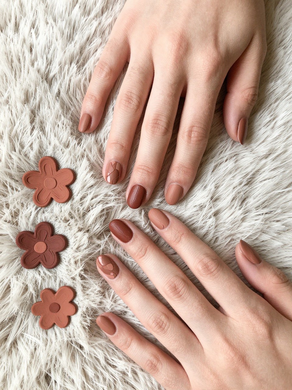 Overhead view of one hands on a fur rug. The nails are a matte clay color. Abstract, painterly floral shapes in a darker terracotta brown adorn each nail. The aesthetic is organic and warm.
