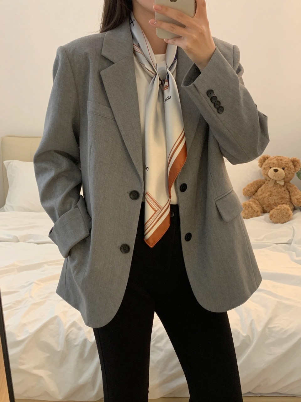 iPhone mirror selfie of a woman in an oversized gray blazer over a white tee and black skinny trousers with a long silk scarf draped at her neck, standing with phone near her shoulder, cropped at NECK so face doesn't show, in a feminine-themed bedroom, evening golden hour, iPhone photo quality.