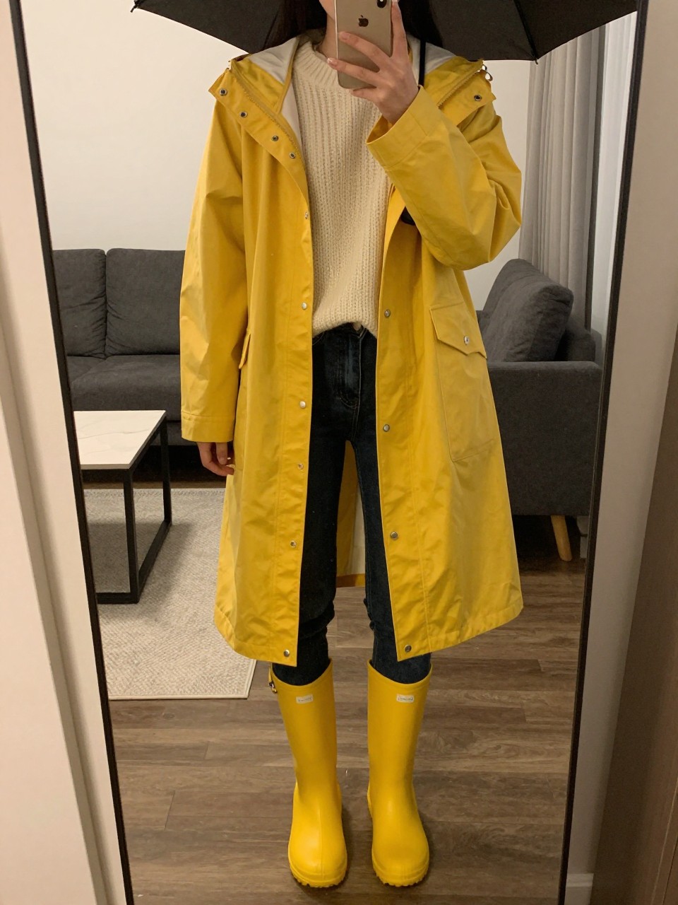 iPhone mirror selfie of a woman in a yellow raincoat over a cream chunky jumper with dark skinny jeans tucked into bright wellies, holding an umbrella, cropped at NECK so face doesn't show, in a city apartment, warm moody lighting, iPhone photo quality.