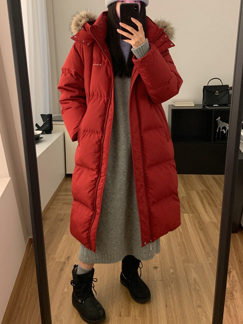 iPhone mirror selfie of a woman wearing the long red puffer coat over an oversized grey sweater dress with black snow boots and a pom-pom beanie, posed with her hands in her coat pockets, cropped at NECK so face doesn't show, in a city apartment with warm moody lighting, iPhone photo quality.