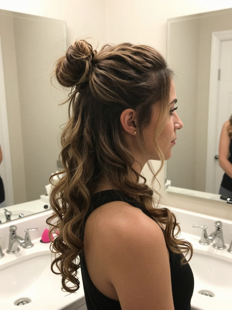 Photo of a woman wearing a textured top knot with long pieces, side profile view showing the knot and the hanging curled pieces, setting: a bathroom with a vanity and mirror, lighting: bright, even indoor lighting, containing a makeup sponge resting on the counter, iPhone photo quality.