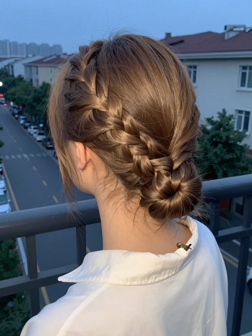 Photo of a white teen girl wearing a rope twist half-up, back-of-head view showing the twists meeting, setting: a balcony overlooking a city street at dusk, lighting: soft, fading natural light, containing a small hair clip clipped to her collar, iPhone photo quality.
