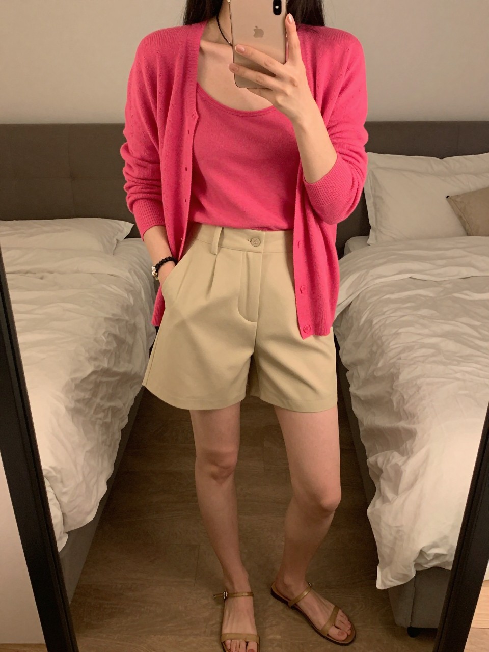 iPhone mirror selfie of a woman in a bright fuchsia cardigan worn as a top with beige tailored shorts and flat sandals, one hand in her pocket, phone in the other, cropped at NECK so face doesn't show, in a feminine-themed bedroom, warm moody lighting, iPhone photo quality.