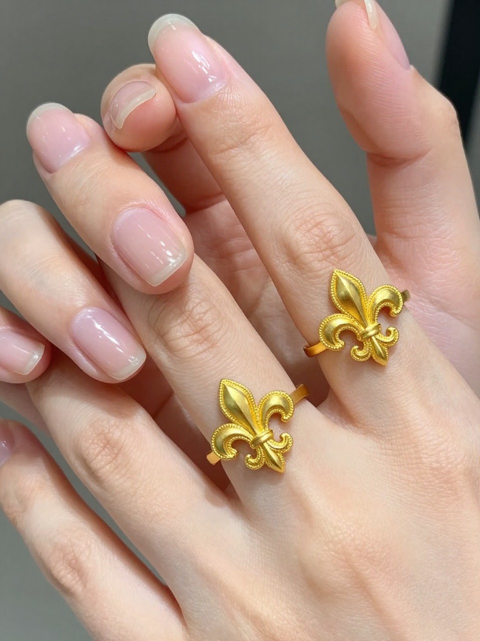 Close-up view of hands near a piece of intricate gold filigree jewelry. The natural, short nails are glossy and clear. A detailed, metallic gold Florentine lily emblem is affixed to each ring finger. The light catches the gold detailing.