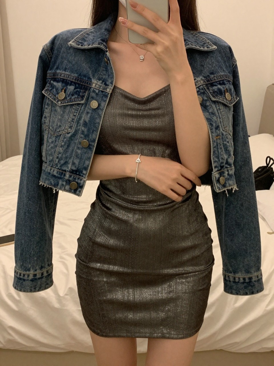 iPhone mirror selfie of a woman in a metallic dress with a denim jacket on her shoulders, adjusting her bracelet, cropped at NECK so face doesn't show, in a feminine-themed bedroom, warm moody lighting, iPhone photo quality.