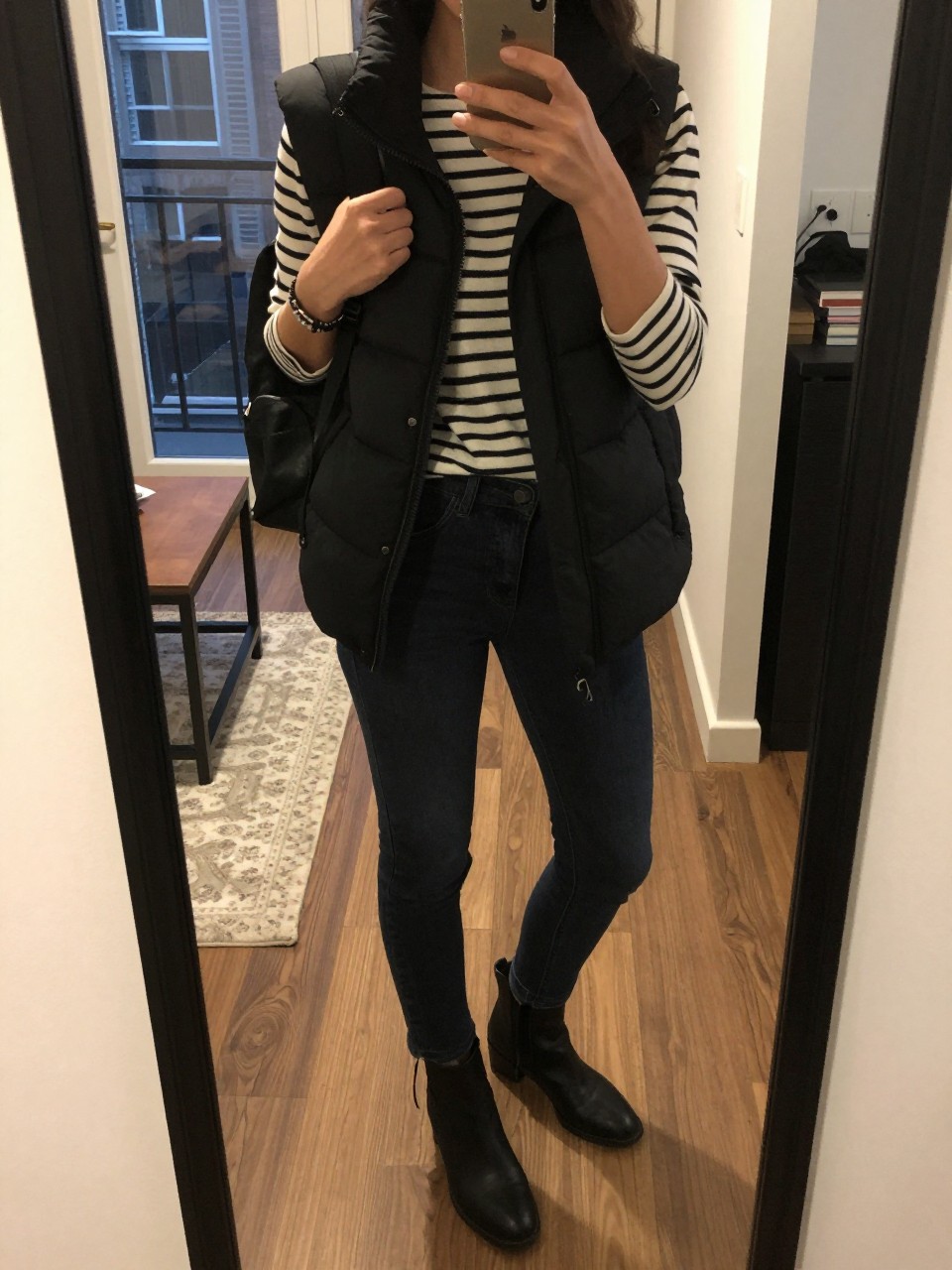 iPhone mirror selfie of a woman in a black puffer vest over a Breton top with dark jeans and ankle boots, adjusting a backpack strap, cropped at NECK so face doesn't show, in a city apartment, warm moody lighting, iPhone photo quality.