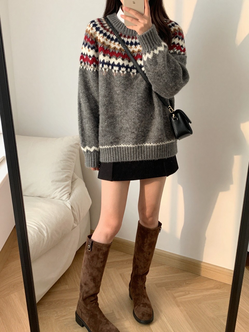 iPhone mirror selfie of a woman wearing the oversized fair isle sweater with a black mini skirt and brown knee-high suede boots, holding a crossbody bag, posed with one leg slightly bent, cropped at NECK so face doesn't show, in a feminine-themed bedroom, evening golden hour, iPhone photo quality.
