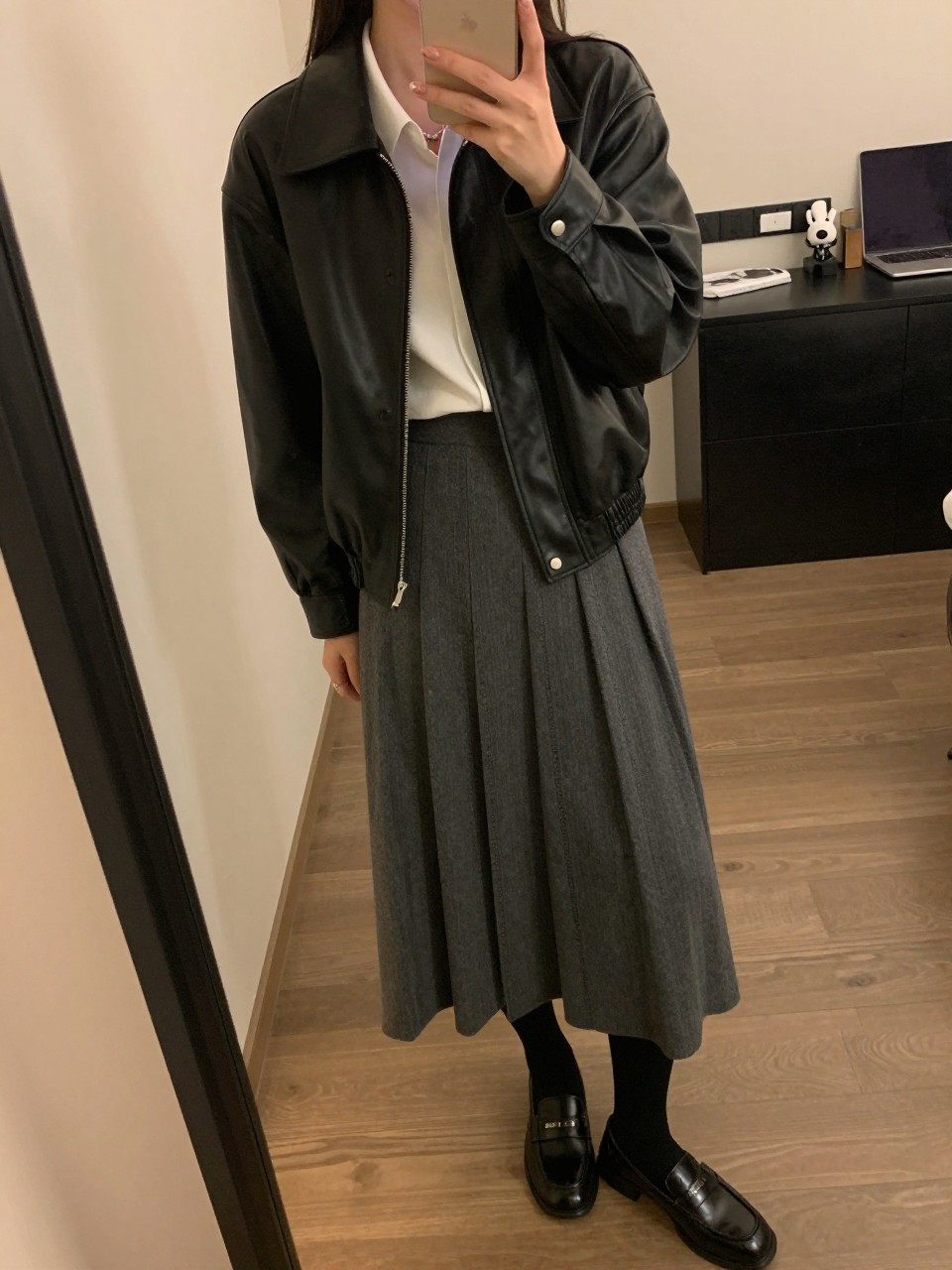iPhone mirror selfie of a woman in a black leather jacket over a white blouse and gray pleated midi skirt with black tights and loafers, standing with phone angled slightly down, cropped at NECK so face doesn't show, in a city apartment, warm moody lighting, iPhone photo quality.