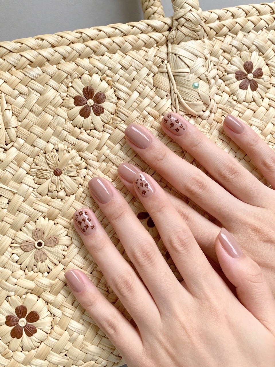 Bird's-eye view of hands next to a woven straw bag with a floral pattern. The nails are a soft mocha. A woven texture with integrated tiny flowers is painted in a contrasting brown on each nail. The detail is intricate.