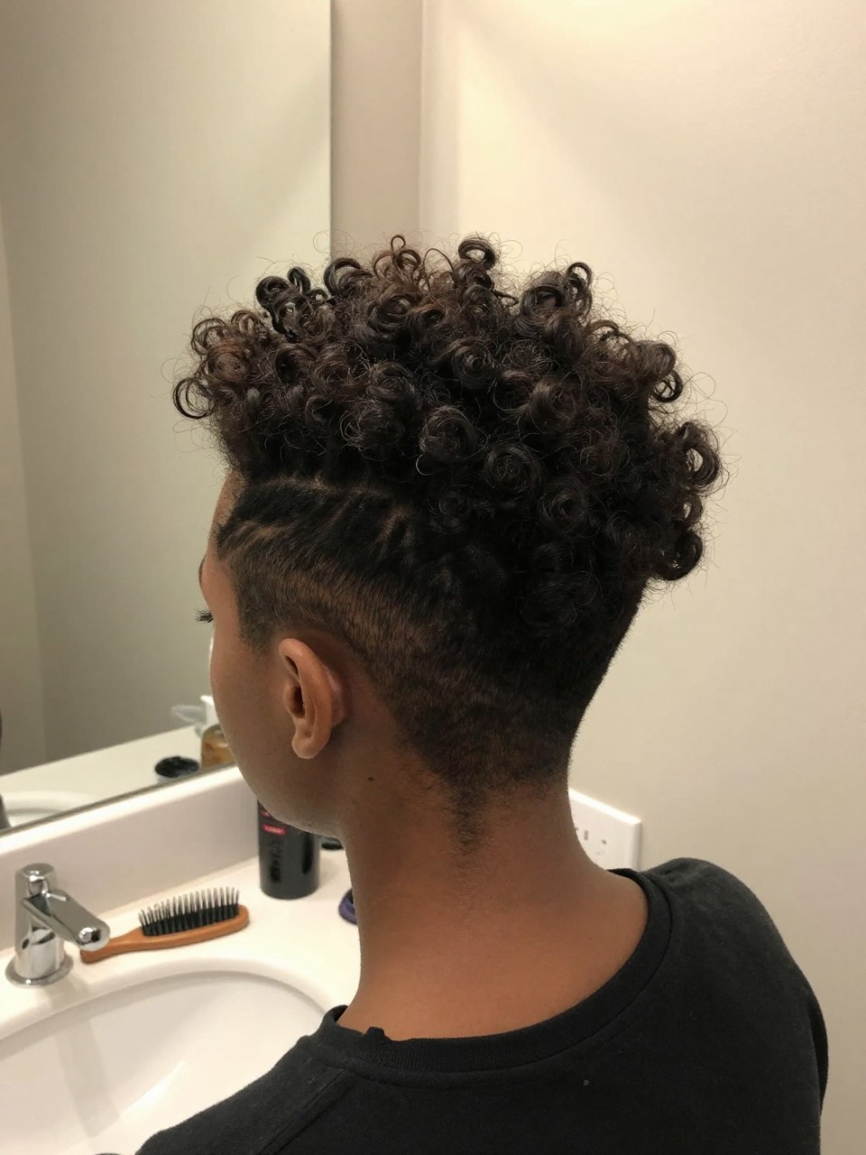 Photo of a black girl wearing a curly mohawk updo, back-of-head view showing the pinned sides and voluminous center strip, in a bathroom with a mirror and soft indoor lighting, containing a rattail comb resting on the counter, iPhone photo quality.