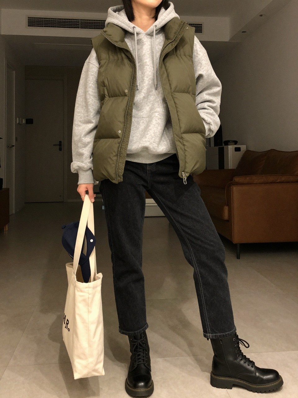 full photo of a woman wearing the olive puffer vest over a heather grey hoodie with dark straight-leg jeans and combat boots, holding a canvas tote and a baseball cap, posed casually with her weight on one leg, cropped at NECK so face doesn't show, in a city apartment with warm moody lighting, iPhone photo quality.