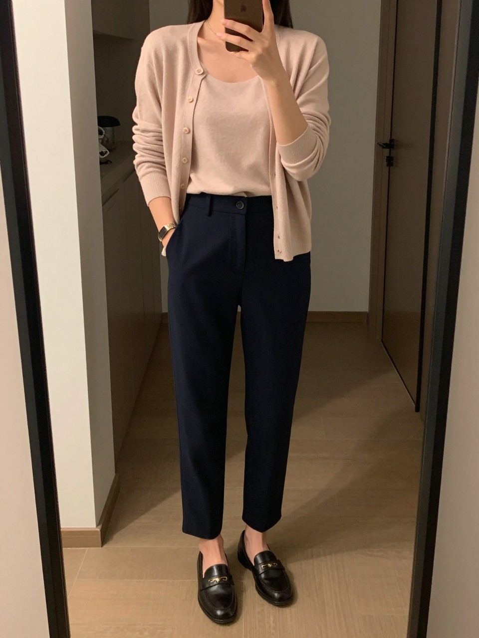iPhone mirror selfie of a woman in a blush cardigan worn as a top tucked into navy tailored trousers with loafers, standing with one hand in her pocket, phone in the other, cropped at NECK so face doesn't show, in a city apartment, warm moody lighting, iPhone photo quality.