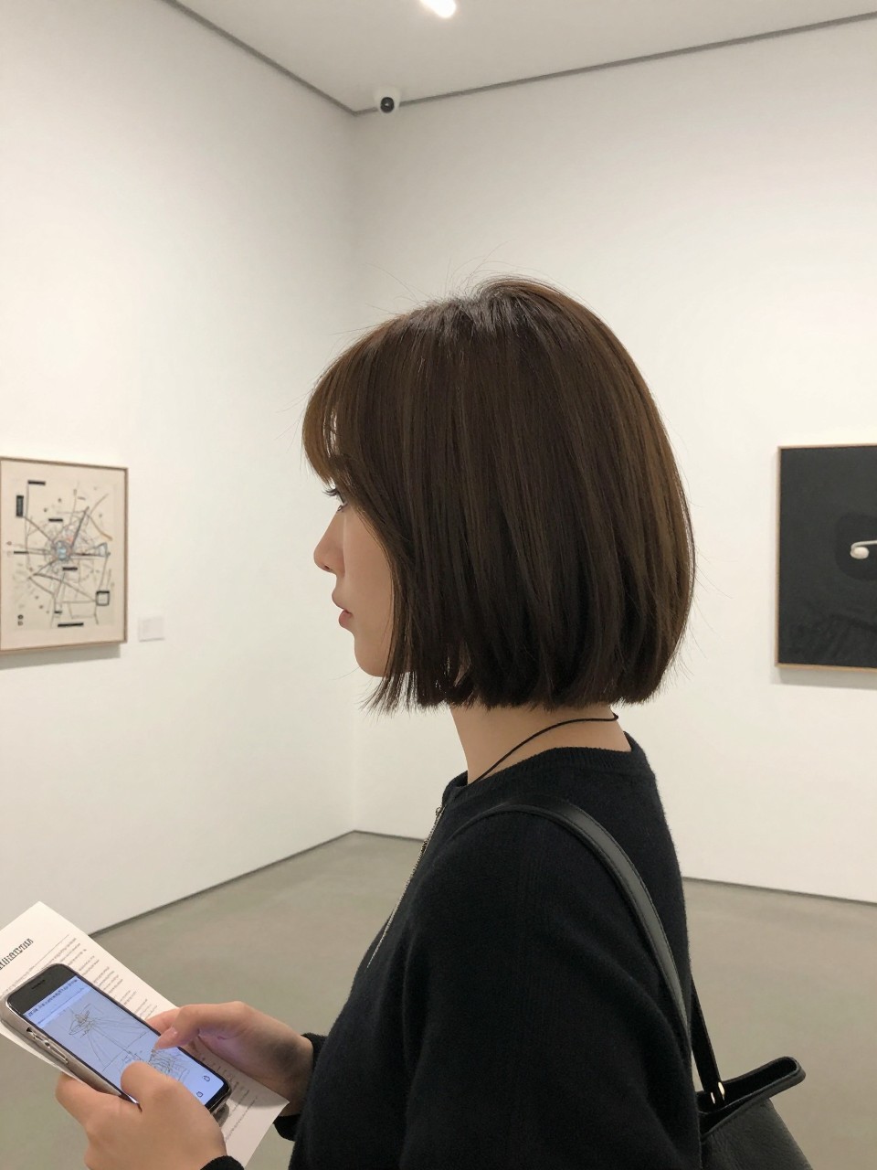 Photo of sleek center-part choppy bob, looking away from camera profile shot, in a modern art gallery with white walls, soft diffused gallery lighting, containing a museum map in hand, iPhone photo quality.