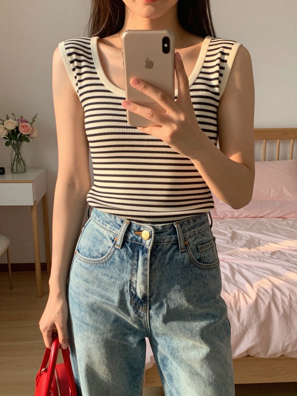 iPhone mirror selfie of a woman in a striped top and jeans, holding a red bag at her side, phone held at chest level, cropped at NECK so face doesn't show, in a feminine-themed bedroom, evening golden hour, iPhone photo quality.