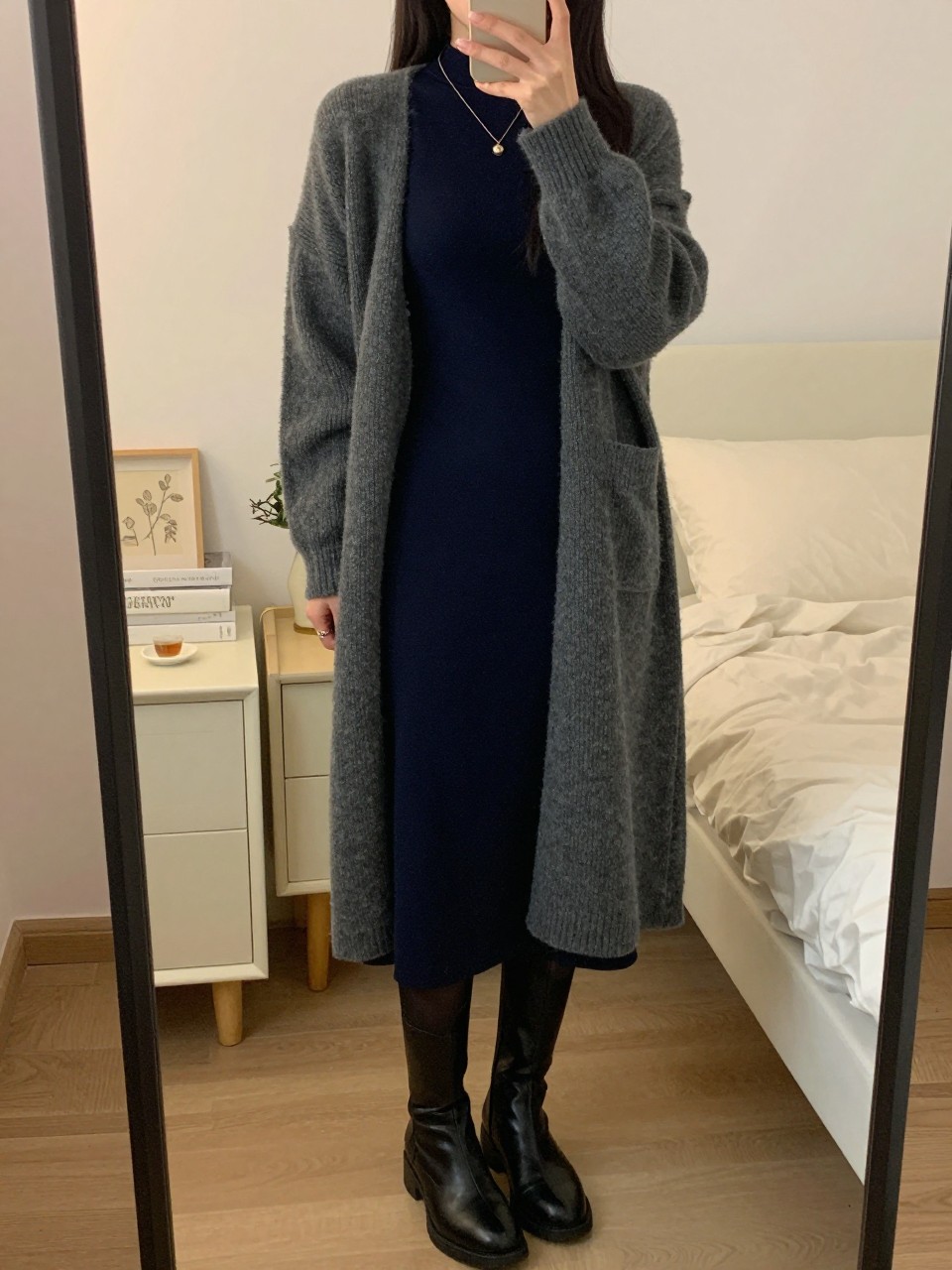 iPhone mirror selfie of a woman in a long gray cardigan over a navy midi dress with black tights and knee-high boots, holding a cup of tea, cropped at NECK so face doesn't show, in a feminine-themed bedroom, warm moody lighting, iPhone photo quality.