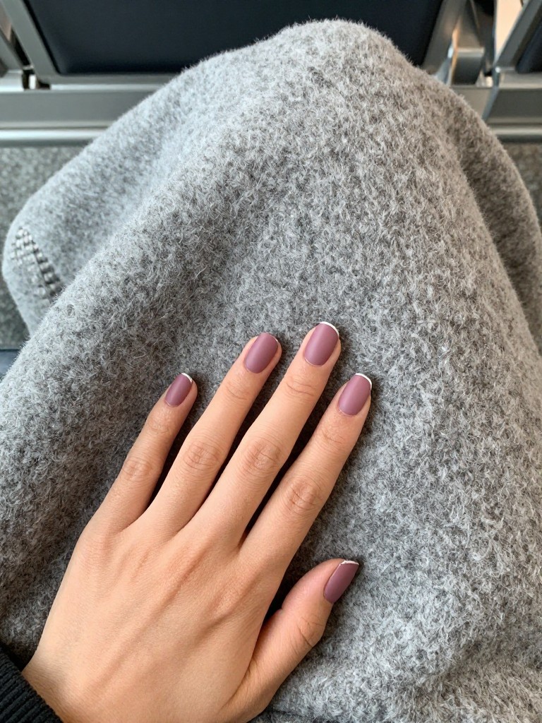  Top-down view of hands on a grey cashmere travel blanket in an airport lounge. The nails are oval in a matte mauve-pink. A thin silver line accents each tip. The lighting is calm and neutral.