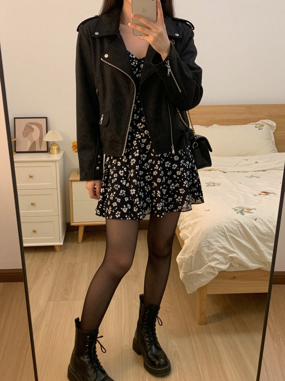 iPhone mirror selfie of a woman in a black moto jacket over a dark floral dress with black tights and combat boots, standing with phone angled down, cropped at NECK so face doesn't show, in a feminine-themed bedroom, evening golden hour, iPhone photo quality.