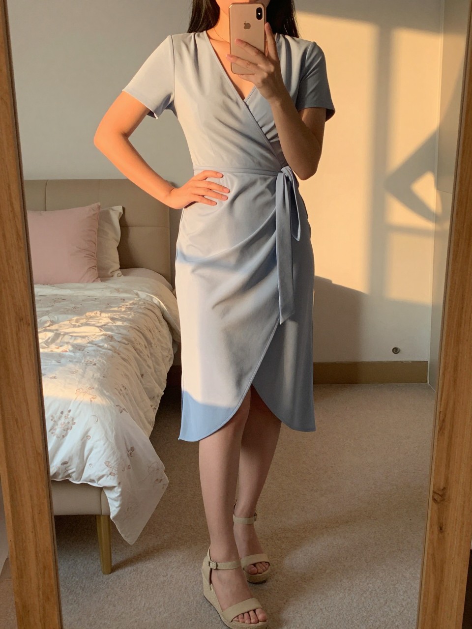 iPhone mirror selfie of a woman in a wrap dress and wedges, holding her phone at waist level, weight shifted to one hip, cropped at NECK so face doesn't show, in a feminine-themed bedroom, evening golden hour, iPhone photo quality.