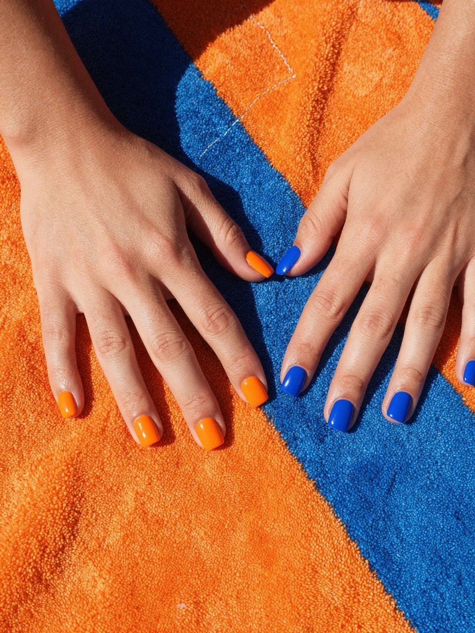 Overhead view of hands on a colorful beach towel with orange and blue stripes. The square nails are diagonally split between glossy orange and cobalt blue. The colors pop under the sun.