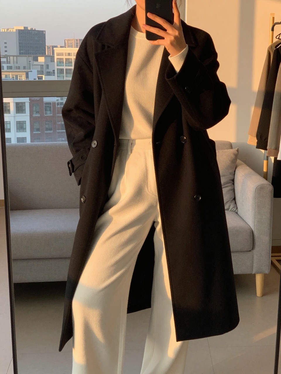 iPhone mirror selfie of a  woman wearing a black duster coat over a cream monochrome outfit, posed with her weight back and phone low, cropped at NECK so face doesn't show, in a city apartment, evening golden hour light, iPhone photo quality.