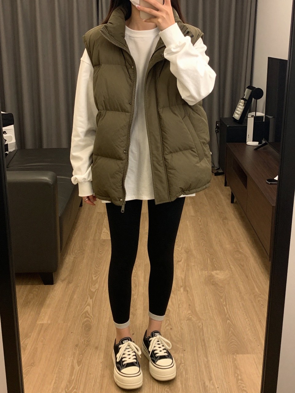 iPhone mirror selfie of a woman in an olive puffer vest over a white long-sleeve tee with black leggings and platform sneakers, standing with phone near her waist, cropped at NECK so face doesn't show, in a city apartment, warm moody lighting, iPhone photo quality.
