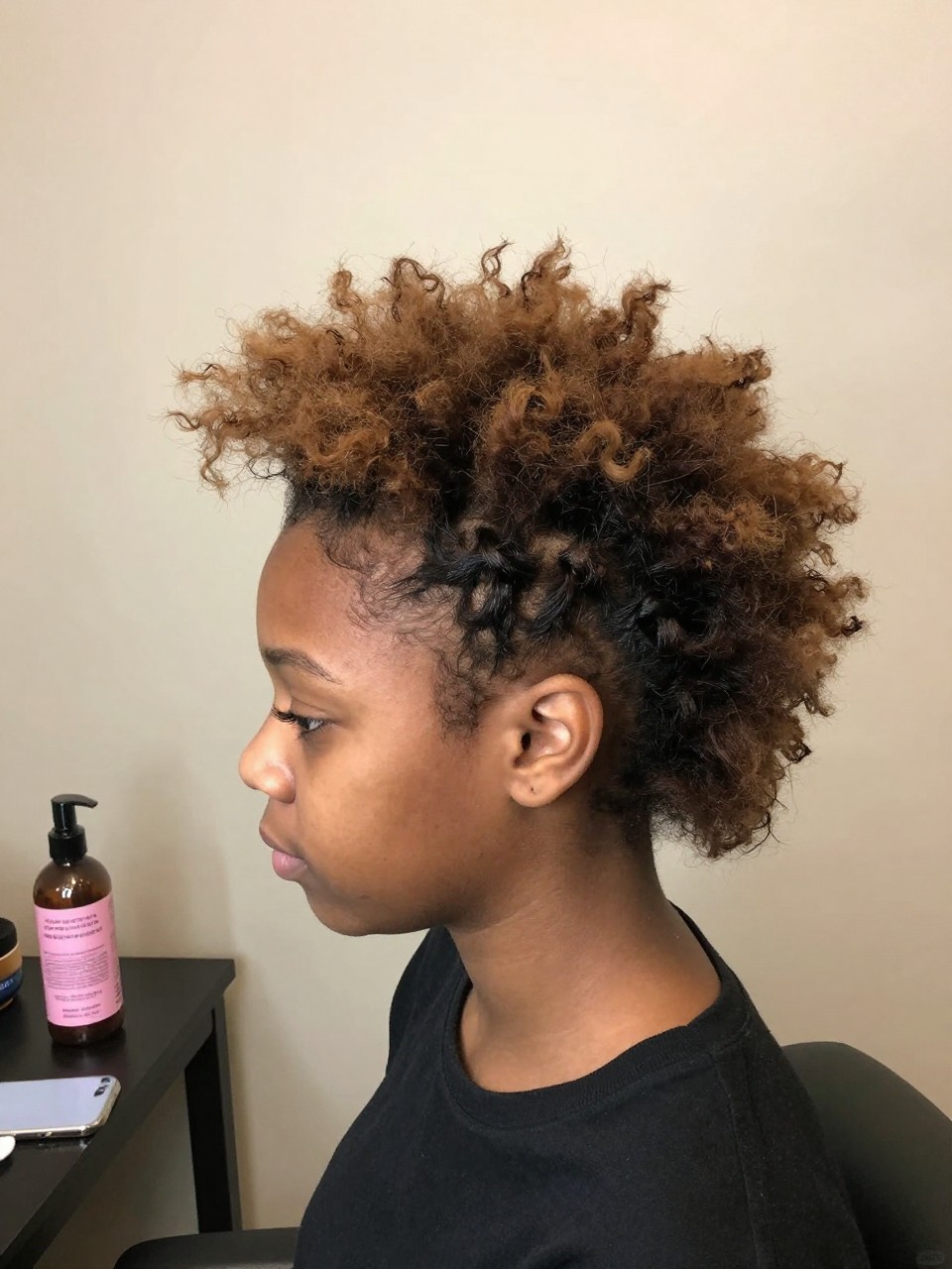 Photo of a black girl wearing a fluffy faux hawk, side profile view showing the pinned side and voluminous center, in soft indoor lighting against a plain wall, containing a bottle of volumizing powder on the table, iPhone photo quality.