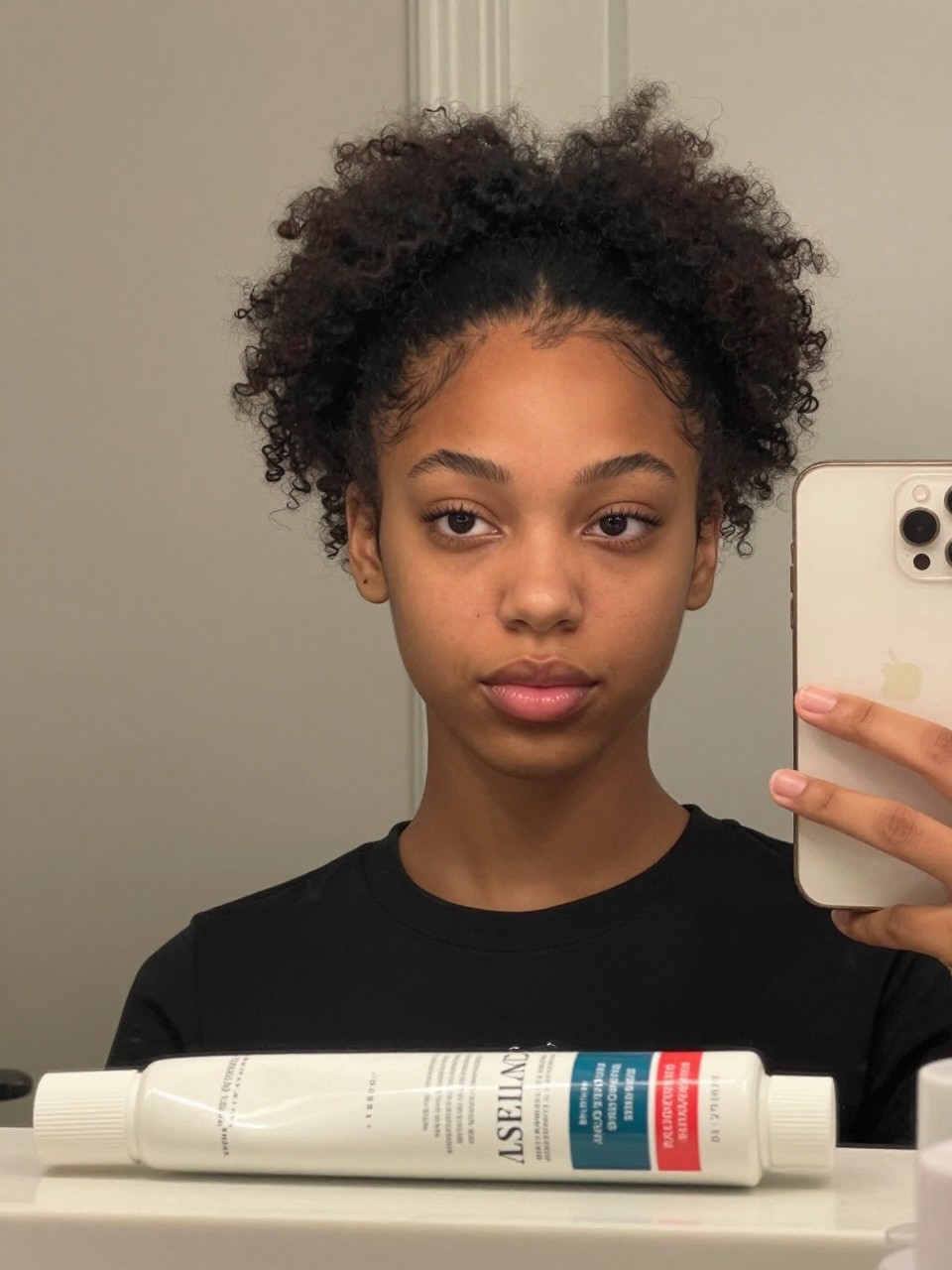 Photo of a black girl wearing a high curly pony with face-framing tendrils, front-facing candid mirror selfie showing the height and soft pieces around the face, in soft indoor lighting, containing a tube of curl cream on the counter, iPhone photo quality.