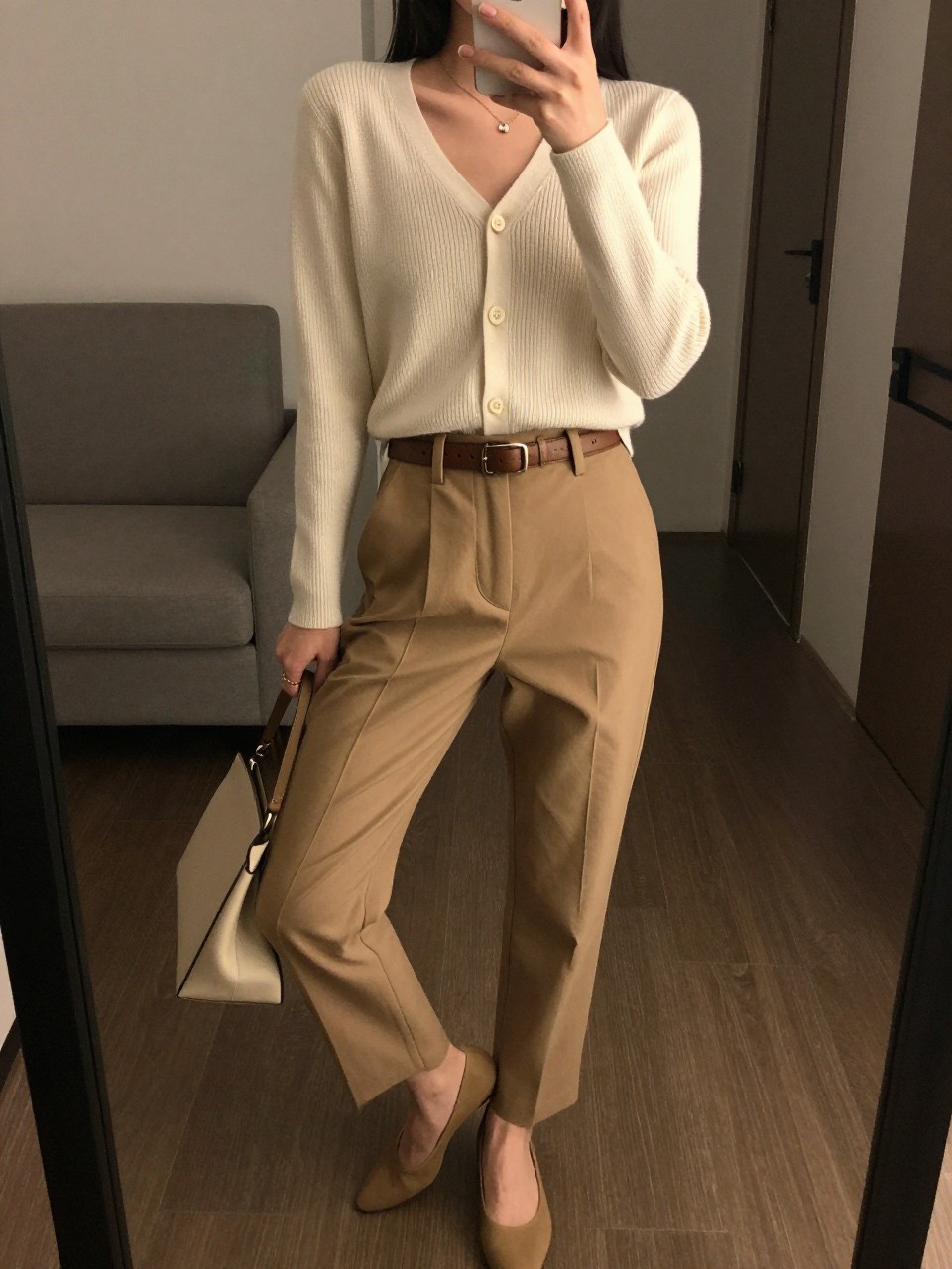 iPhone mirror selfie of a woman wearing a cream fine-gauge cardigan buttoned as a top with camel tailored trousers cinched with a leather belt and pointed-toe flats, holding a structured shoulder bag, posed with her weight on one leg, cropped at NECK so face doesn't show, in a city apartment, warm moody lighting, iPhone photo quality.