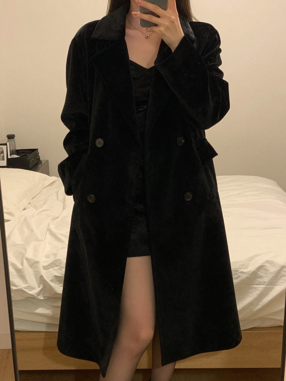 iPhone mirror selfie of a woman wearing a black velvet coat over formal evening wear, standing with her phone low and body angled gracefully, cropped at NECK so face doesn't show, in a feminine-themed bedroom, warm moody lighting, iPhone photo quality.