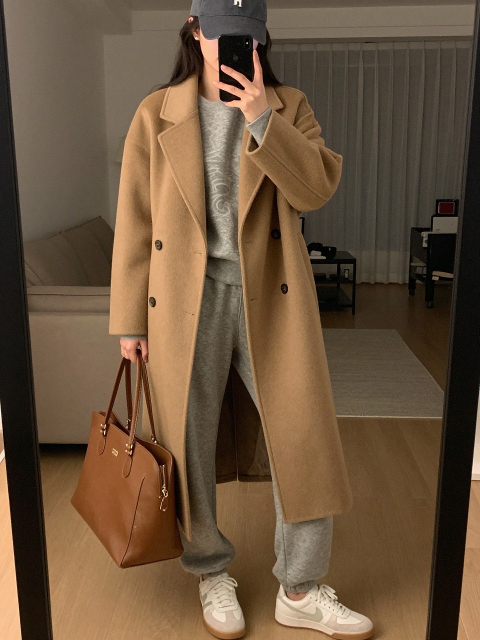 iPhone mirror selfie of a woman wearing the camel wool coat over a matching grey sweatsuit with white sneakers and a baseball cap, holding a leather tote, posed casually with her weight back, cropped at NECK so face doesn't show, in a city apartment, warm moody lighting, iPhone photo quality.