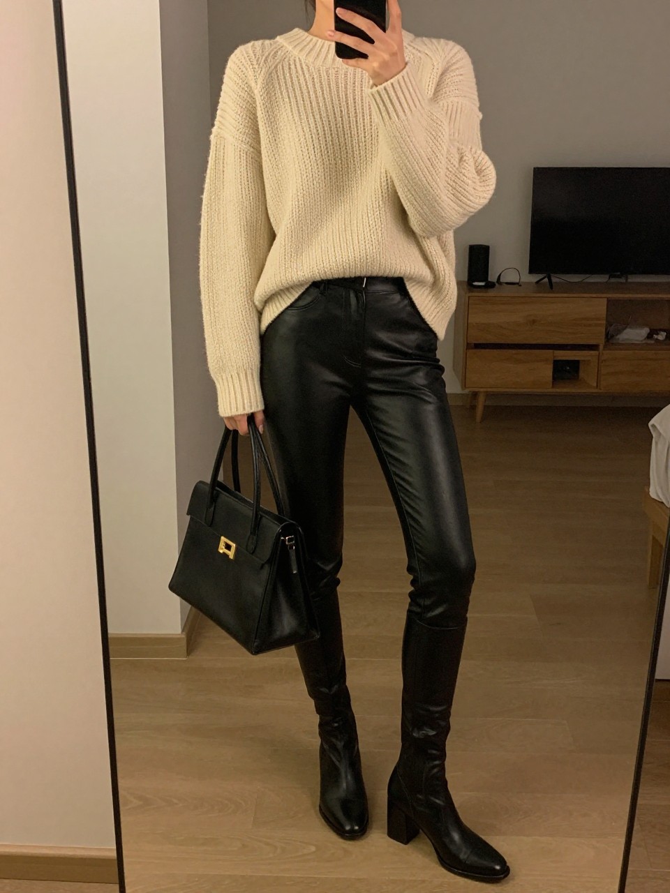iPhone mirror selfie of a woman wearing the cream chunky sweater tucked into black leather leggings and block-heel boots, holding a structured black bag, posed with one foot slightly forward, cropped at NECK so face doesn't show, in a city apartment, warm moody lighting, iPhone photo quality.