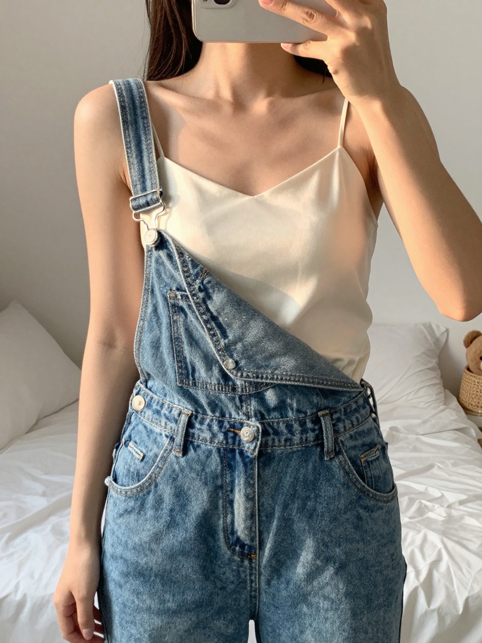 iPhone mirror selfie of a woman in denim overalls over a satin cami, hooking one overall strap, cropped at NECK so face doesn't show, in a feminine-themed bedroom, golden hour sunlight, iPhone photo quality.