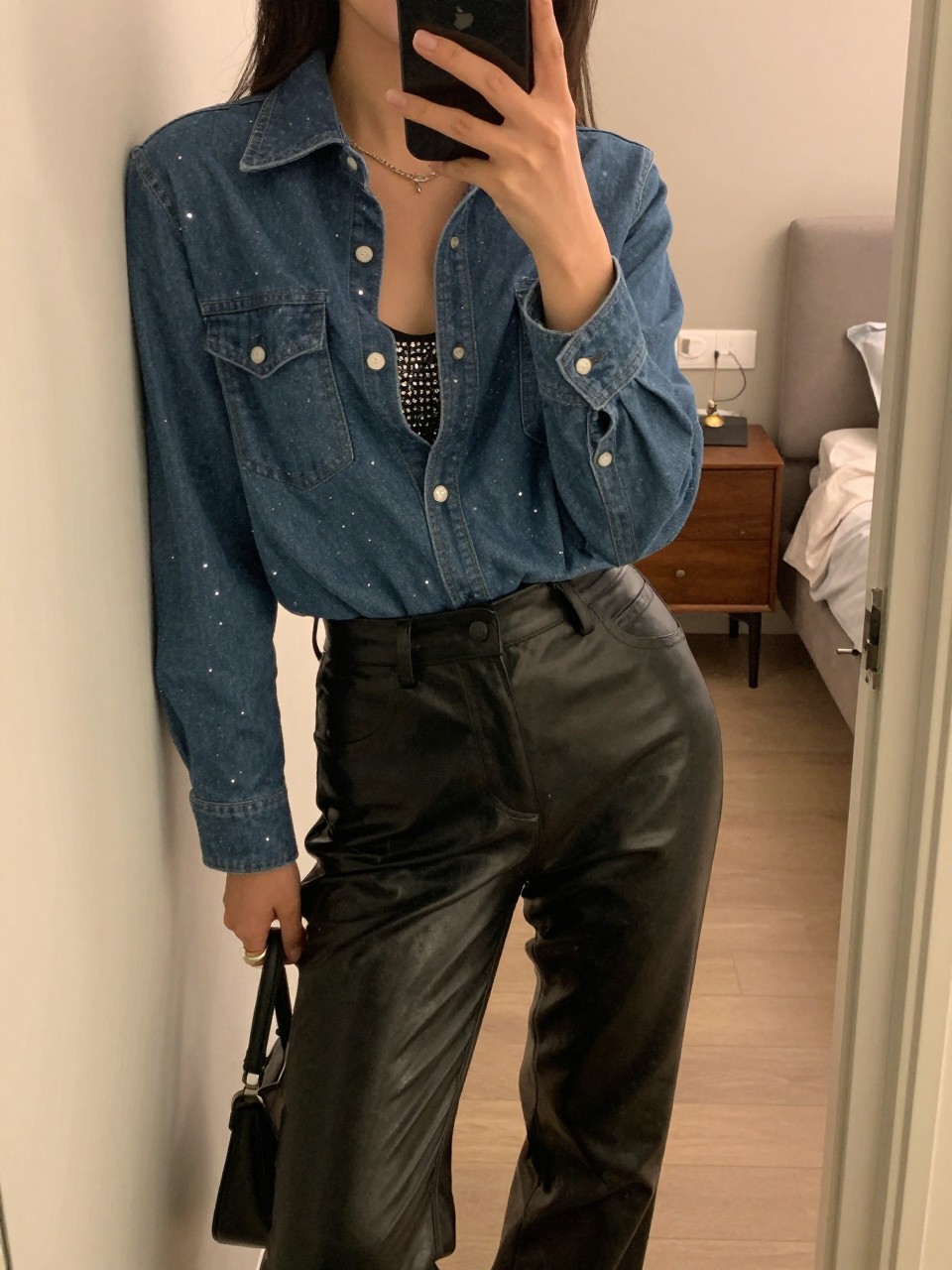 iPhone mirror selfie of a woman in a sparkly denim shirt over leather pants and a sequin cami, leaning against a door frame, cropped at NECK so face doesn't show, in a city apartment, warm moody lighting, iPhone photo quality.