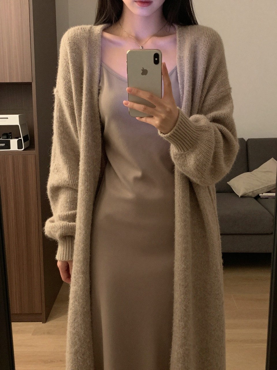 iPhone mirror selfie of a woman in a long cardigan over a tee and slip skirt, holding her phone at waist level, looking down at her outfit, cropped at NECK so face doesn't show, in a city apartment, warm moody lighting, iPhone photo quality.