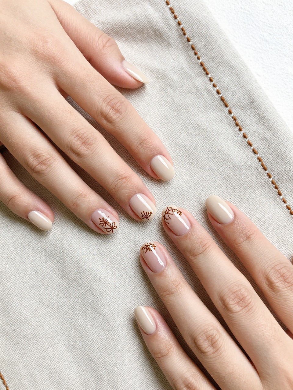 Image Prompt: Overhead shot of two hands on a linen napkin with embroidered details. The nails are a flat beige. Raised, brown gel lines form tiny, intricate floral embroidery patterns on each nail. The texture is clear in the light.