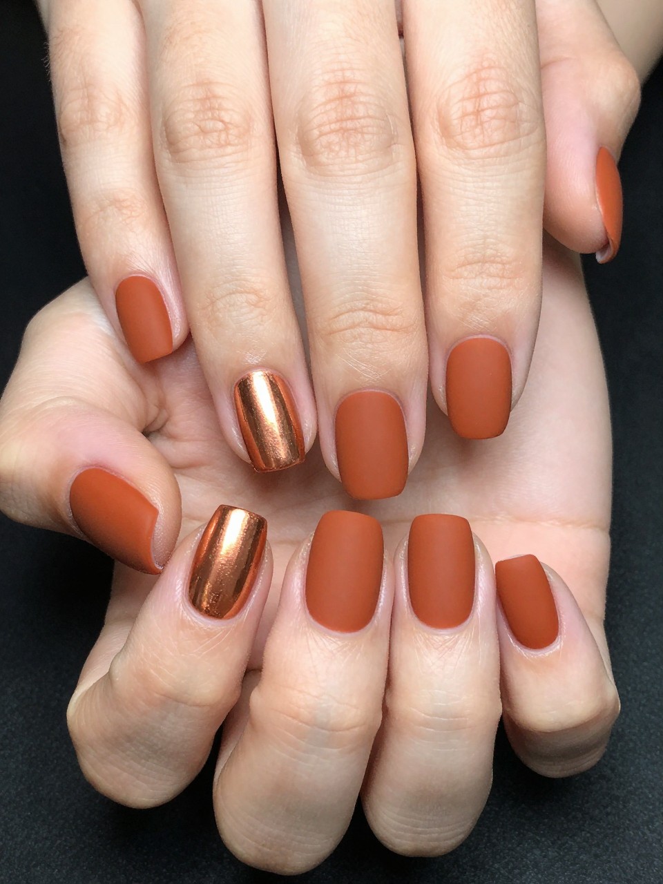 Overhead shot of hands nails are matte rust orange. Each ring finger has a shiny copper stripe. The textures complement each other beautifully.