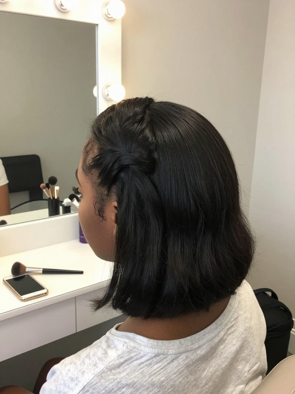 Photo of a teenage black girl wearing a half-up on a bob, back-of-head view showing the gathered section, setting: a bedroom with a vanity and mirror, lighting: soft indoor lighting, containing a makeup brush resting on the vanity, iPhone photo quality.