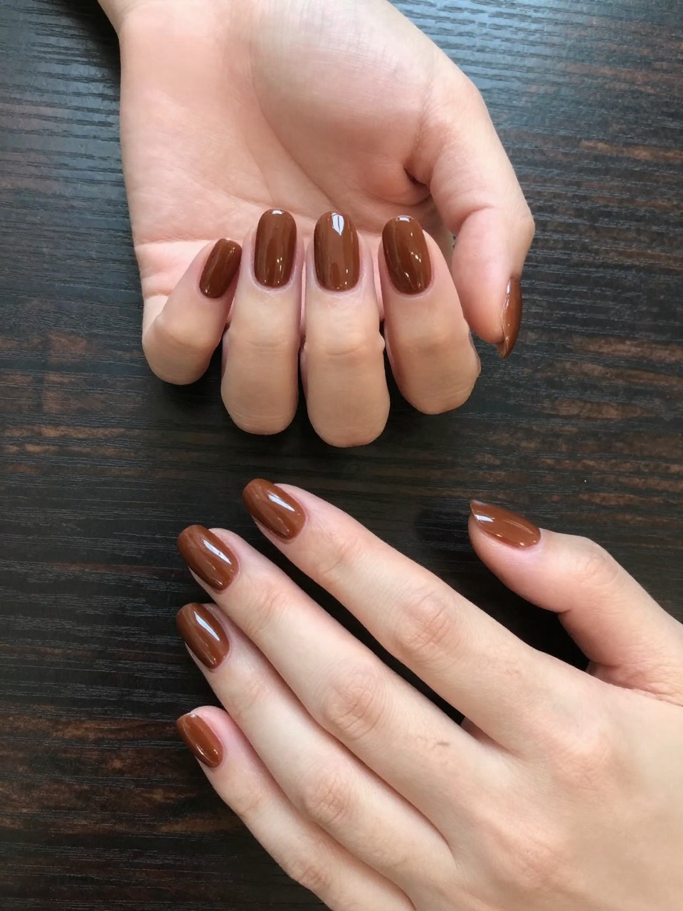Overhead view of hands against a dark wood background. The nails are a high-gloss chestnut brown. The contrast is stunning and elegant.