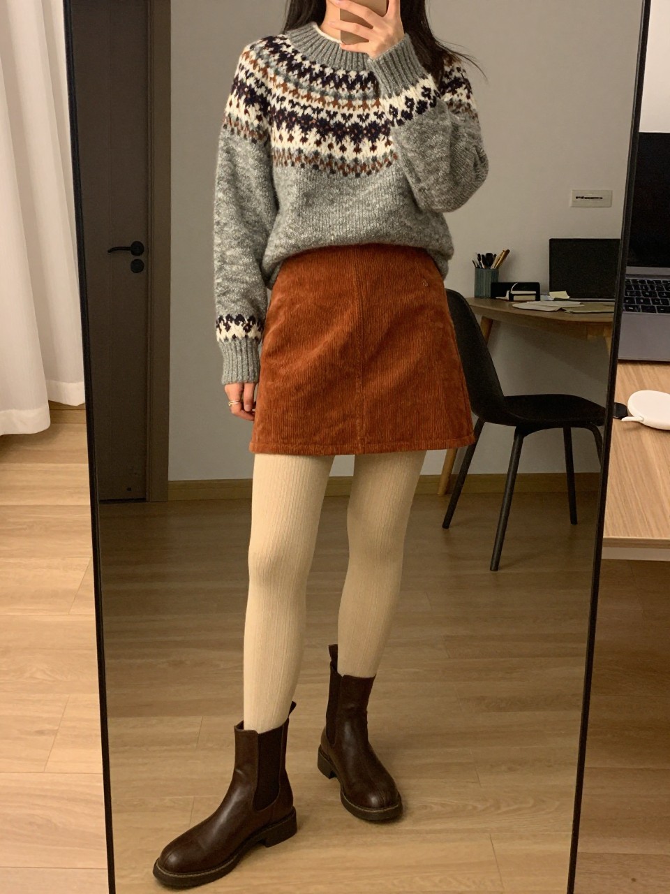 iPhone mirror selfie of a woman in a Fair Isle jumper with a rust corduroy mini skirt, cream tights, and brown Chelsea boots, standing with one foot forward, cropped at NECK so face doesn't show, in a city apartment, warm moody lighting, iPhone photo quality.